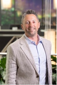 Chris Andrews, Sales Manager at Tindall. He is pictured standing in front of foliage and wearing a gray suit jacket and blue collared shirt.